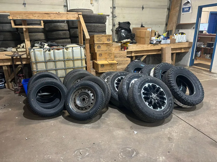 Customer tire sets standing on the shop floor at AutoNorth in North Bay, some mounted on black steel rims and some on alloy wheels, with more tires racked on wooden shelving behind them